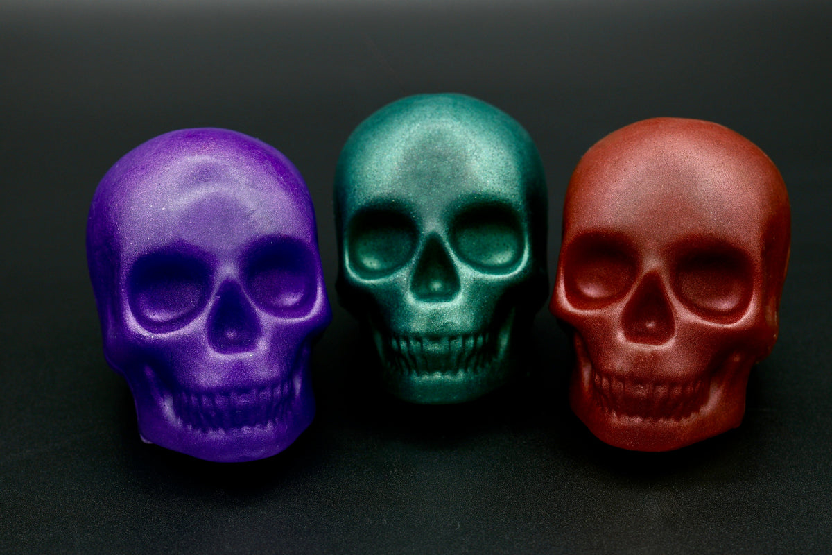 Three skull soaps in purple, teal and red displayed front-facing on dark background